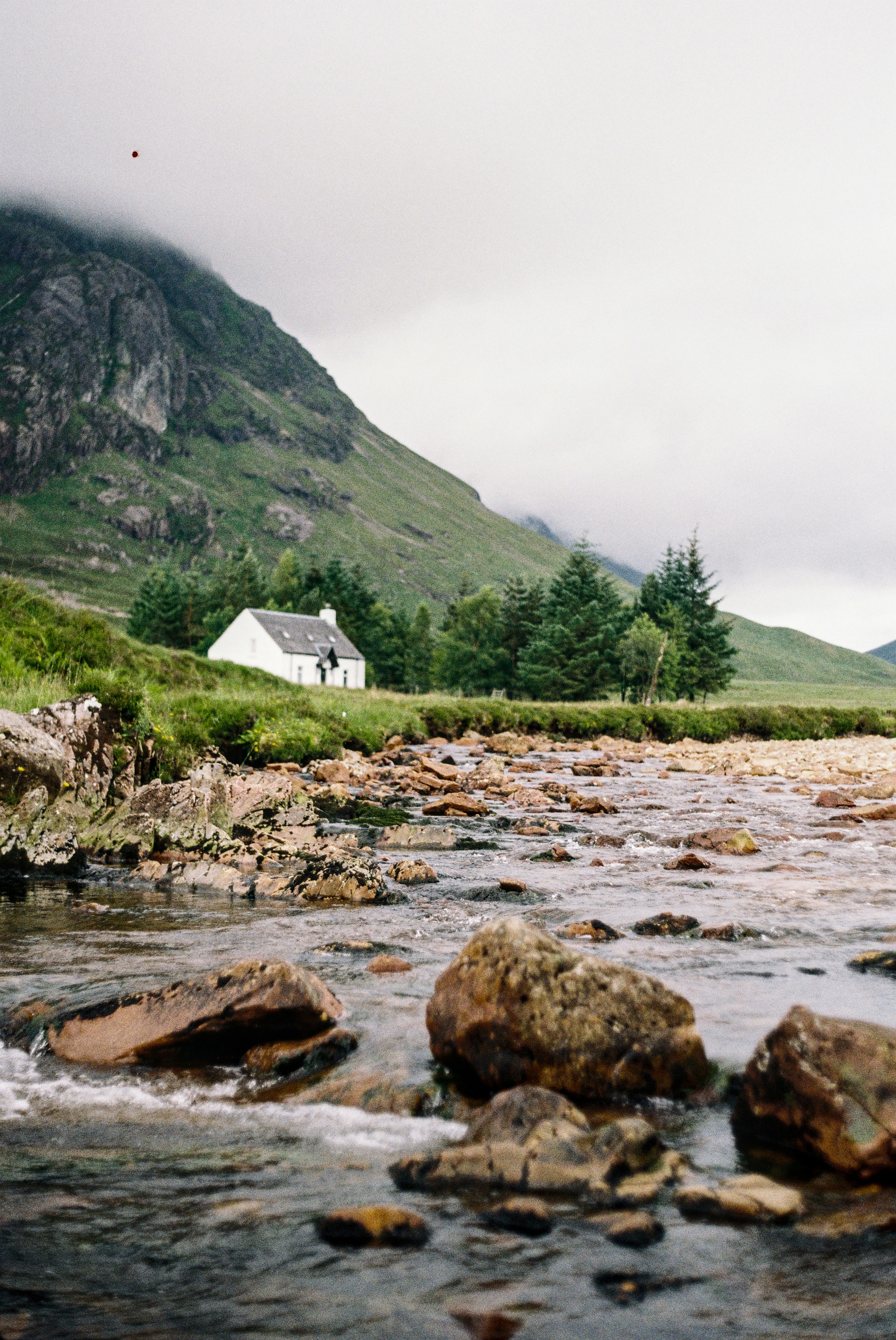 Eine kleine weiße Hütte, die neben einem Bach liegt, umgeben von bewaldeten Bergen